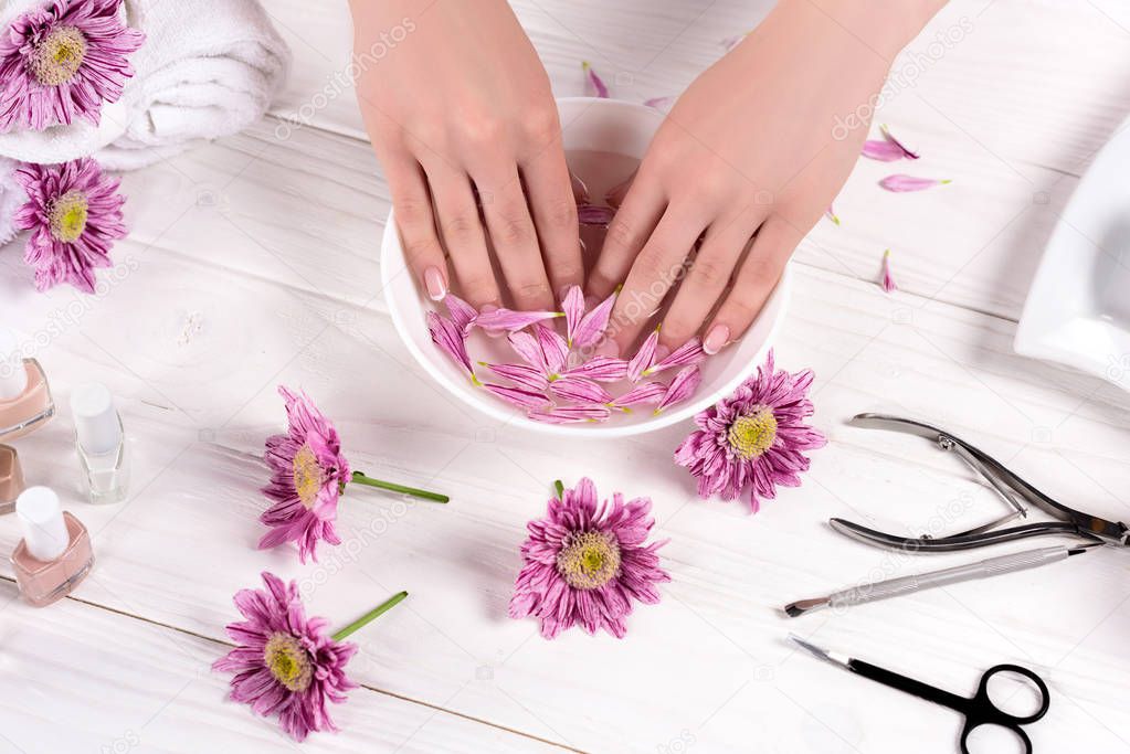 cropped shot of woman receiving bath with petals for nails at table with flowers, towels, nail polishes and tools for manicure in beauty salon 