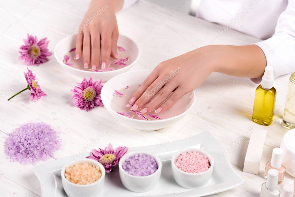 partial view of woman receiving bath for nails at table with flowers, colorful sea salt, cream container, aroma oil bottles and nail polishes in beauty salon 