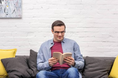mutlu yetişkin adam beyaz tuğla duvar önünde evde kanepede kitap okuma