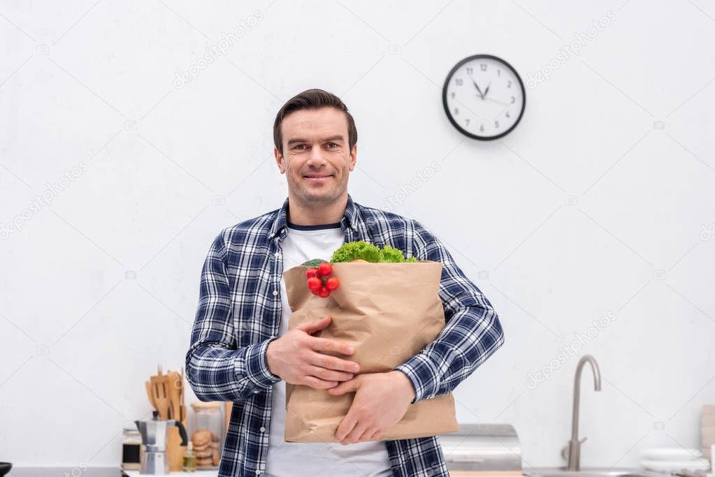 happy adult man carrying grocery store bag at kitchen and looking at camera