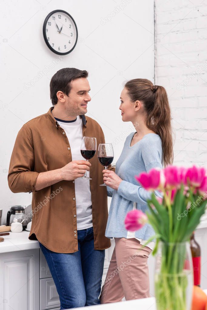 happy adult couple clinking glasses of wine at kitchen with blurred bouquet on foreground