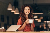 portrét usmívající se žena s šálkem kávy u stolu s notebookem v kavárně