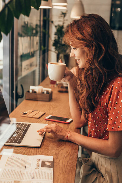 side view of young blogger with cup of coffee working on laptop in coffee shop