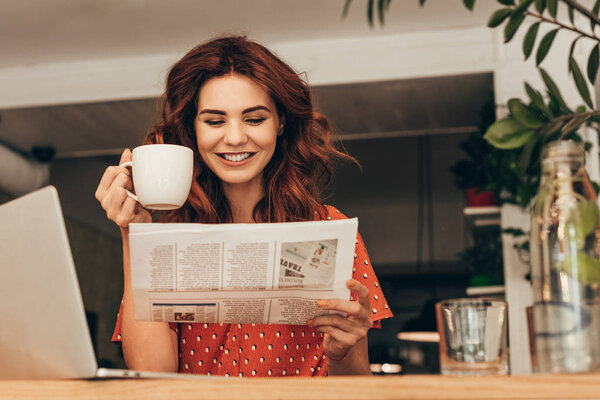 portrait of smiling woman with cup of coffee reading newspaper at table with laptop in coffee shop