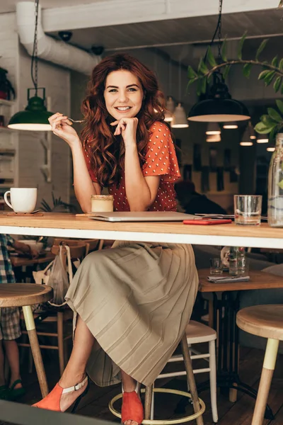 微笑的妇女坐在桌与甜点和笔记本电脑在咖啡馆里 — 图库照片