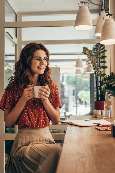 微笑的年轻妇女拿着杯子咖啡在桌与笔记本在咖啡馆 — 图库照片