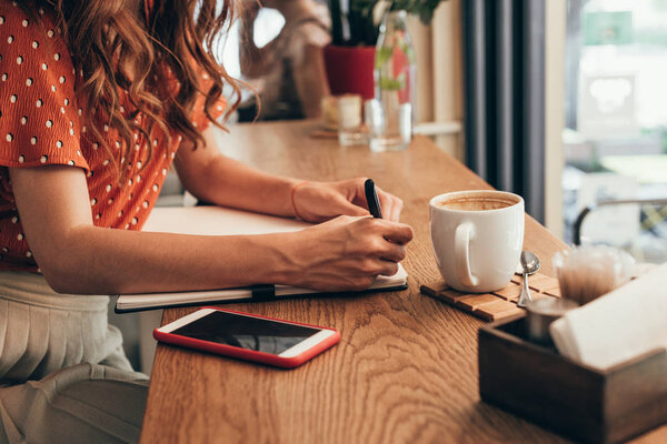обрезанный снимок блогера, делающего заметки в блокноте за столом с чашкой кофе в кафе
