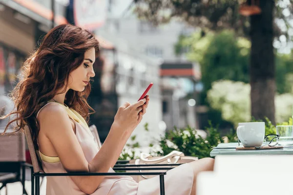 年轻女子使用智能手机的侧面视图坐在桌边咖啡馆里喝咖啡 — 图库照片