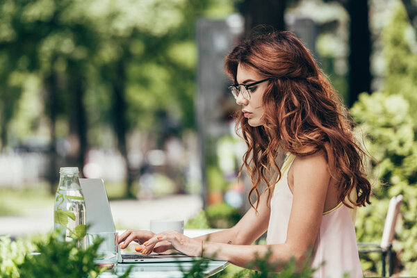 side view of focused young blogger in eyeglasses working on laptop in cafe