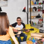 Вид сзади девушек с коробками для пиццы и помпонами, смотрящих на улыбающегося мужчину, играющего на гитаре на домашней вечеринке