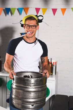 happy young man in beer hat holding keg of beer and smiling at camera clipart