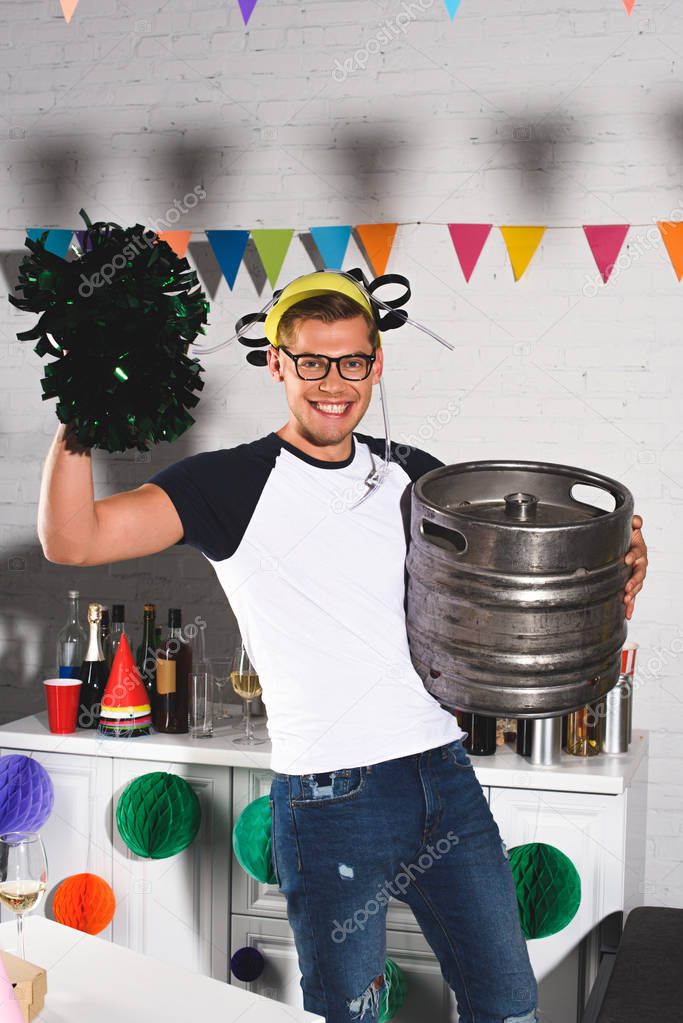cheerful young man in beer hat holding pop-pom and beer barrel at home party