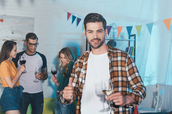 smiling young man holding glass of wine and showing thumb up while partying with friends