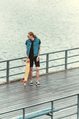 yakışıklı şık dövmeli adamın longboard ile Köprü üzerinde duran yüksek açılı görünüş