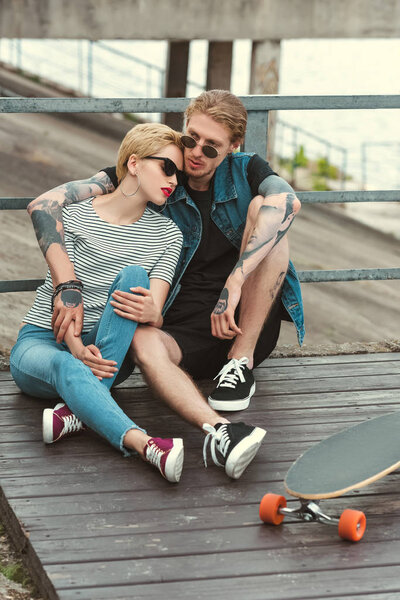 boyfriend with tattoos and stylish girlfriend sitting on bridge and hugging near skateboard