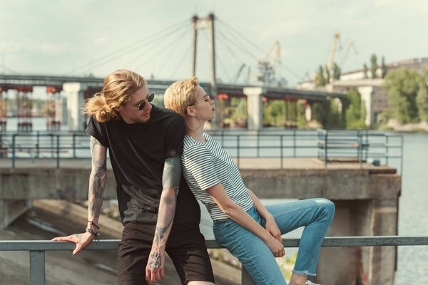 boyfriend with tattoos and stylish girlfriend touching with backs and sitting on railing of bridge