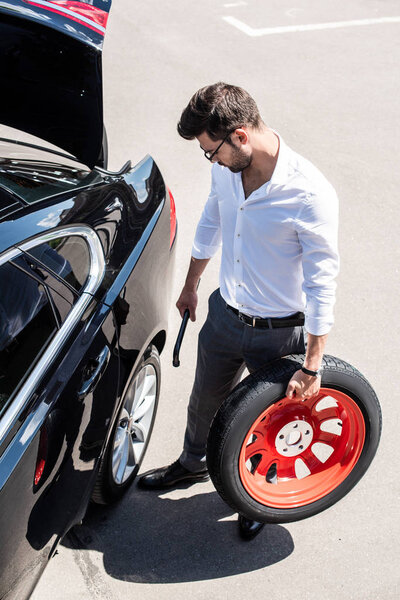 high angle view of young businessman in eyeglasses holding wheel spanner and tire near car at street