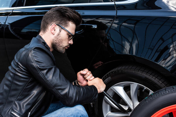 focused young man in sunglasses using wheel spanner for wheel replacement at street