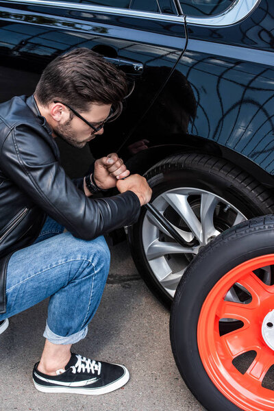 high angle view of young man in sunglasses using wheel spanner for wheel replacement at street