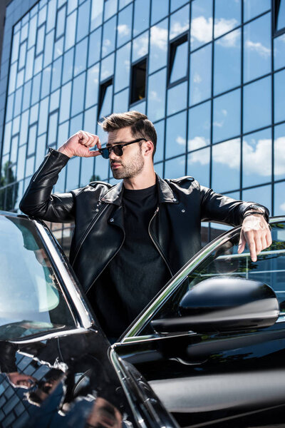 stylish man in leather jacket and sunglasses looking away near car at street