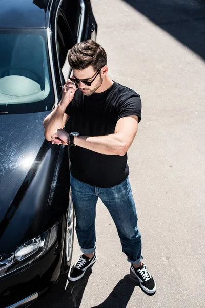 高角度的时尚年轻人在太阳镜检查手表附近他的车在街上 — 免费的图库照片