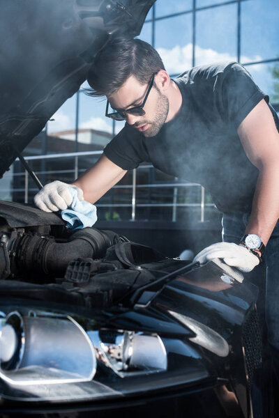 young man in working gloves trying to repair engine of broken car with smoke coming out 