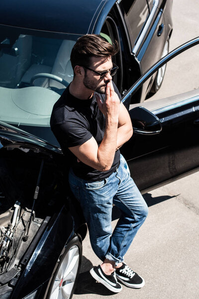 high angle view of stylish man in sunglasses closing mouth by fingers near broken car with opened bonnet