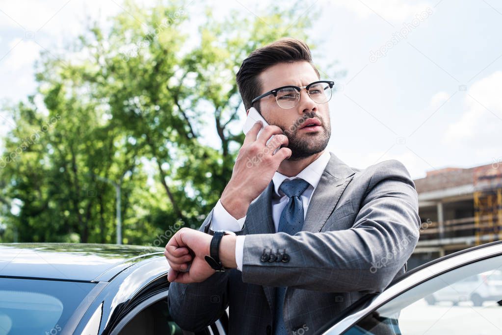 portrait of businessman in eyeglasses talking on smartphone while standing near car on street