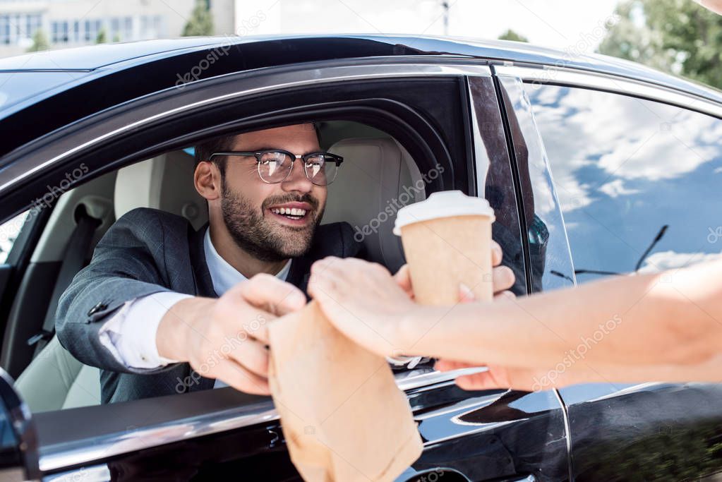 cropped shot of smiling businessman taking coffee to go and take away food from waiter while sitting in car