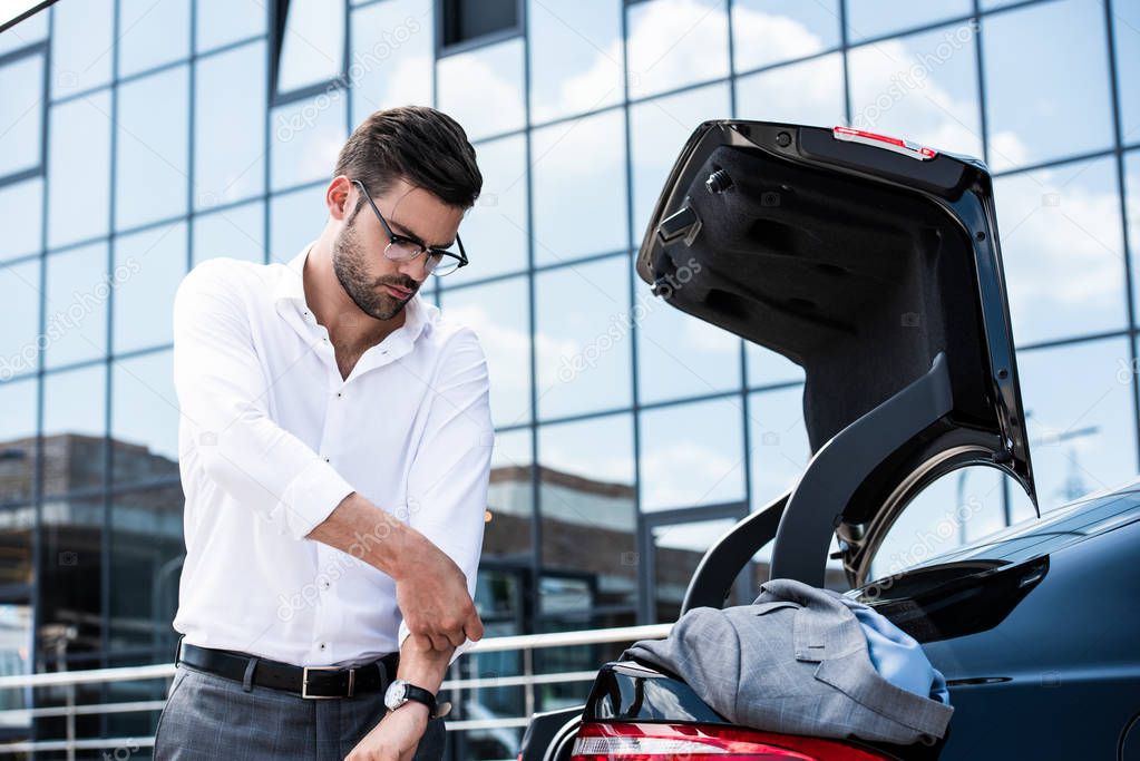 young businessman in eyeglasses rolling sleeves of white shirt near car trunk at street