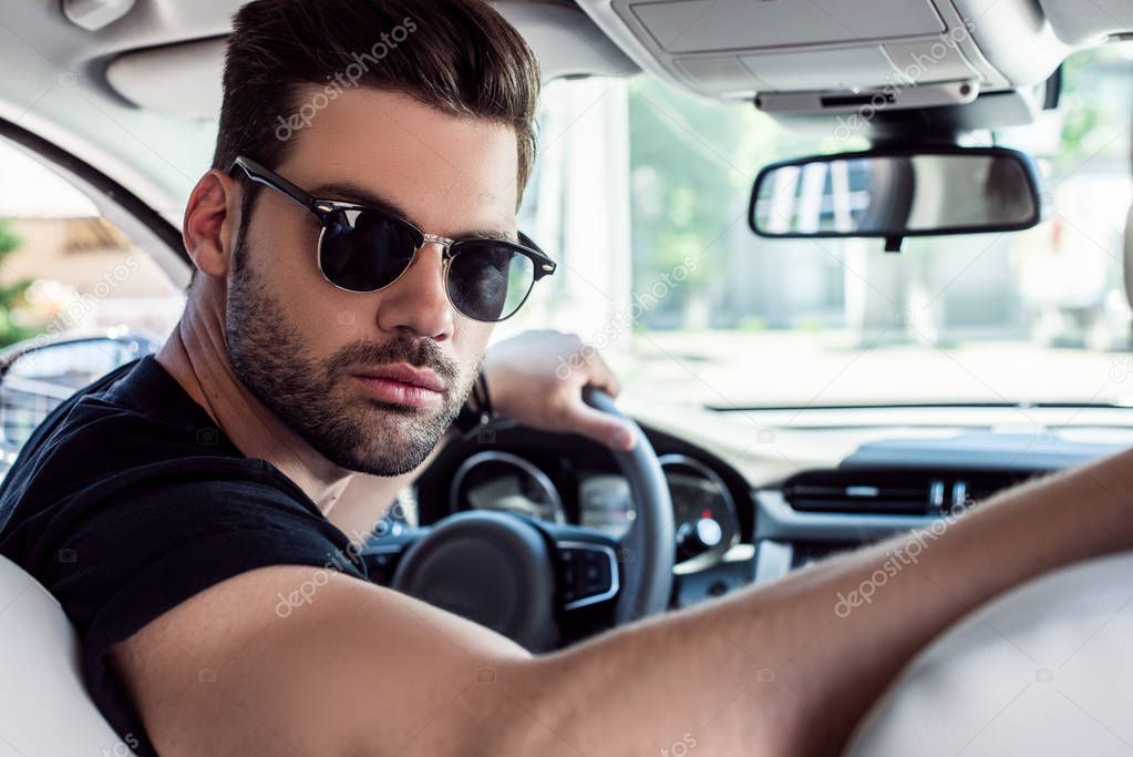 portrait of serious stylish man in sunglasses looking back while driving car 