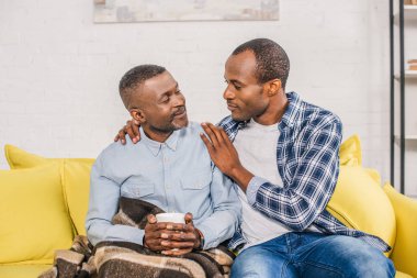 ekose altında oturan ve Evde Yetişkin oğlu seyir sıcak içecek fincan gülümseyen kıdemli adam