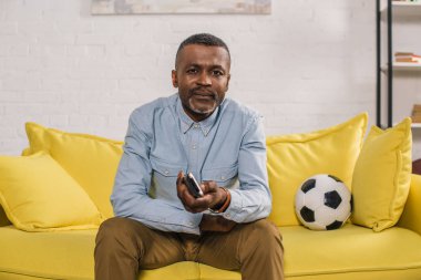 Uzaktan kumanda kullanarak ve futbol topu ile kanepede otururken kameraya bakıyor Afrika kökenli Amerikalı adam