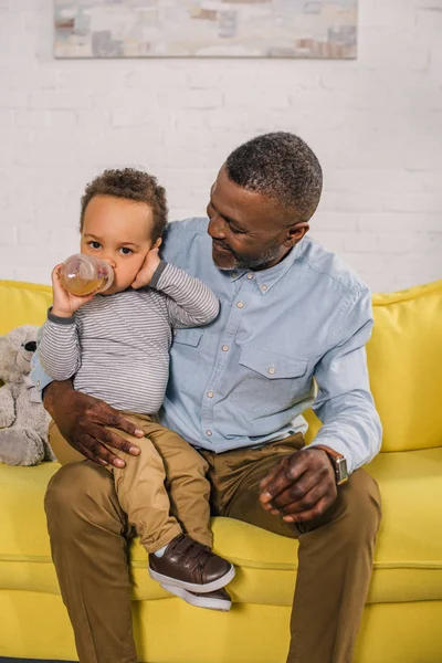 küçük torunu evde bebek şişeden içme bakarak mutlu Afro-Amerikan Büyükbaba 