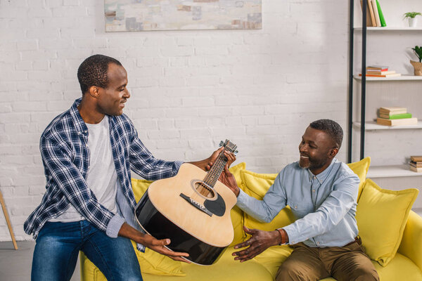 african american father and adult son holding acoustic guitar at home