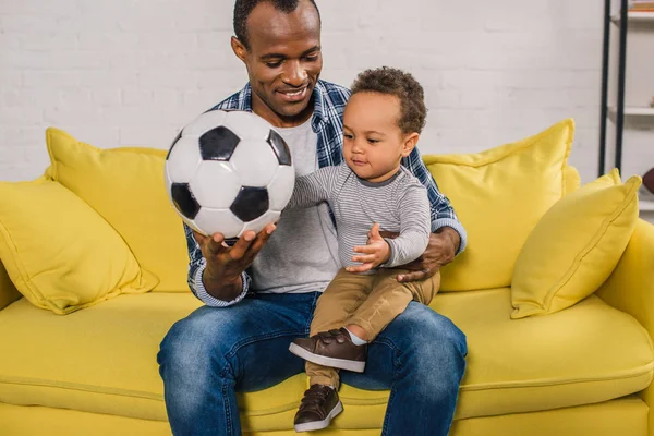 happy young father holding soccer ball while sitting with adorable little son on sofa