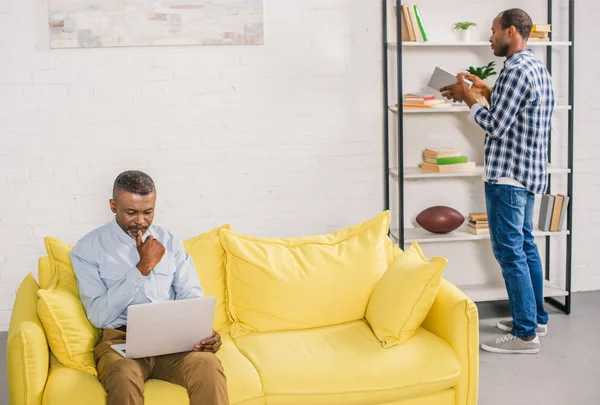 Senior Man Using Laptop While Adult Son Holding Book Bookshelves — Free Stock Photo