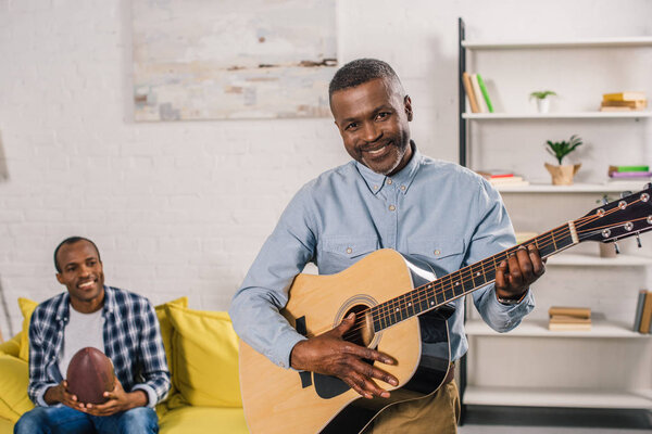 senior man playing acoustic guitar and smiling at camera while adult son holding rugby ball behind