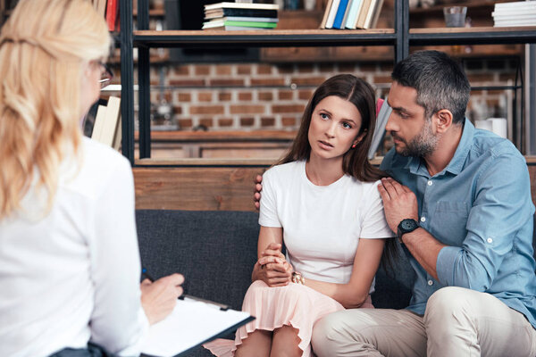 rear view of female psychiatrist taking notes in clipboard while man embracing wife on therapy session in office 