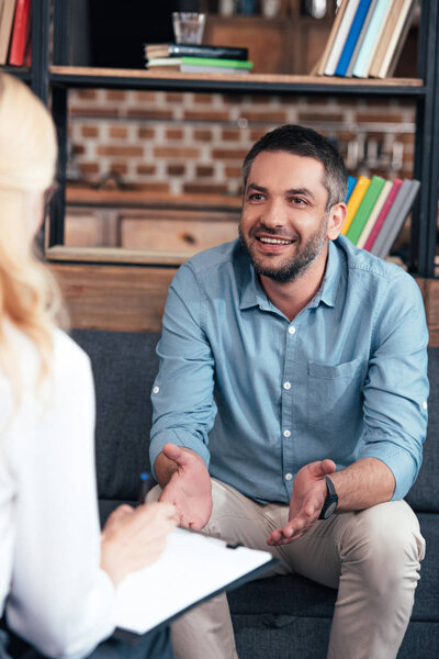 cropped image of female psychiatrist writing in clipboard and talking to smiling man gesturing by hands in office 