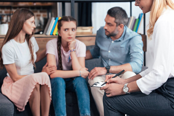 cropped image of female counselor writing in clipboard while parents cheering up daughter on therapy session