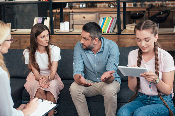 parents gesturing by hands while their daughter using digital tablet on therapy session by female counselor in office 