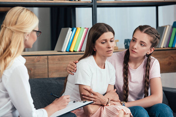 female counselor writing in clipboard while daughter embracing depressed mother on therapy session 