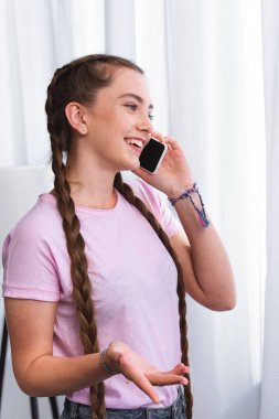 side view of smiling teenage girl with plaits gesturing by hand and talking on smartphone in front of curtains at home clipart