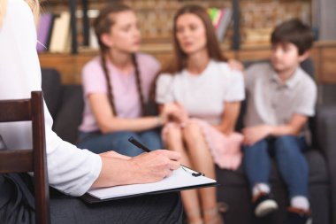 cropped image of female counselor writing in clipboard while children cheering up depressed mother on therapy session  clipart