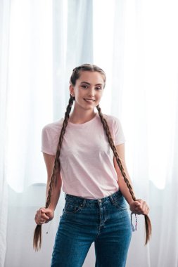 happy teenage girl looking at camera and holding own plaits in front of curtains at home  clipart