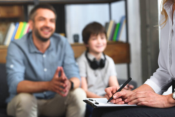 cropped image of female counselor writing in clipboard while father and son sitting on therapy session 