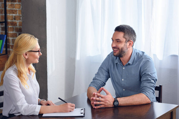 smiling man talking to female psychiatrist while she writing in clipboard at office 
