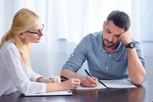 man writing down in empty textbook while sitting at table near female counselor in office 