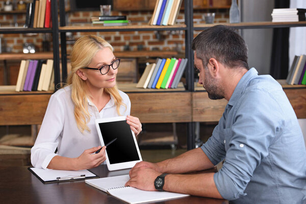 female counselor pointing on digital tablet with blank screen by pen to male patient at table in office 